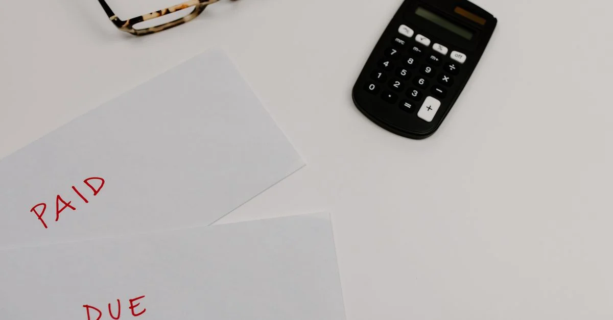 Vue de dessus de papiers financiers étiquetés 'Payé' et 'Dû' à côté d'une calculatrice et de lunettes.