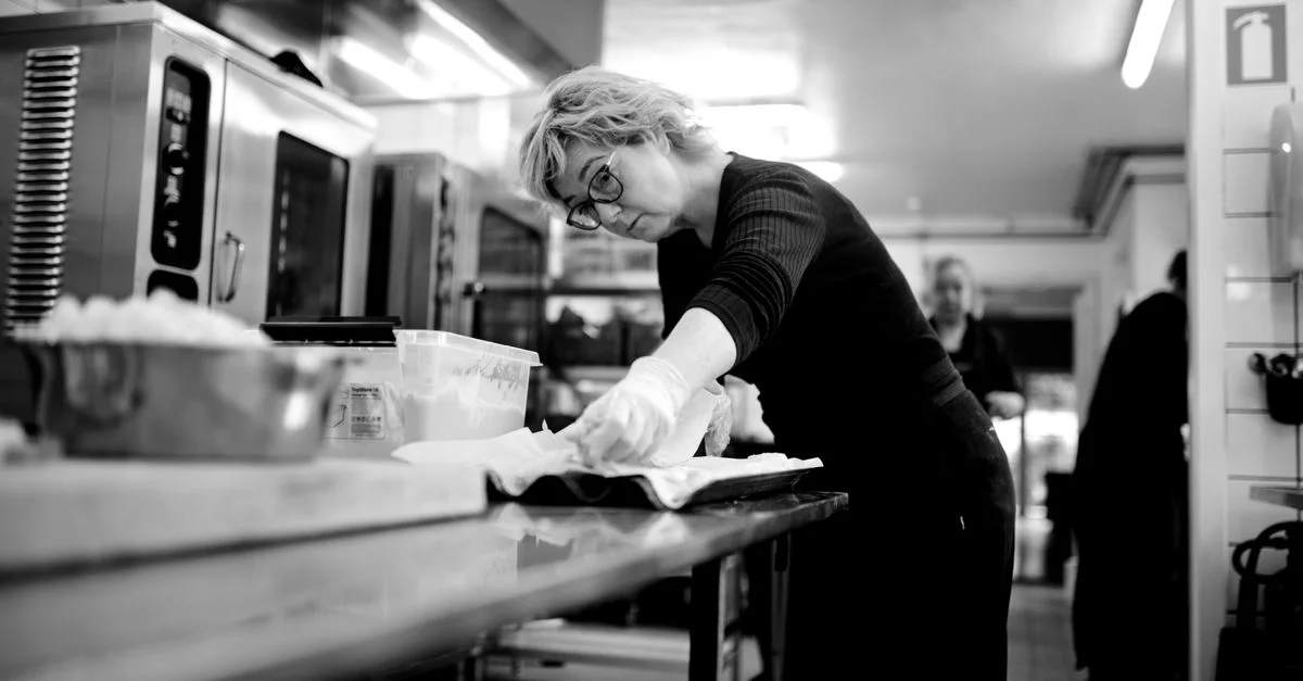 Chef féminine concentrée préparant un plat dans une cuisine commerciale moderne. Image en noir et blanc.